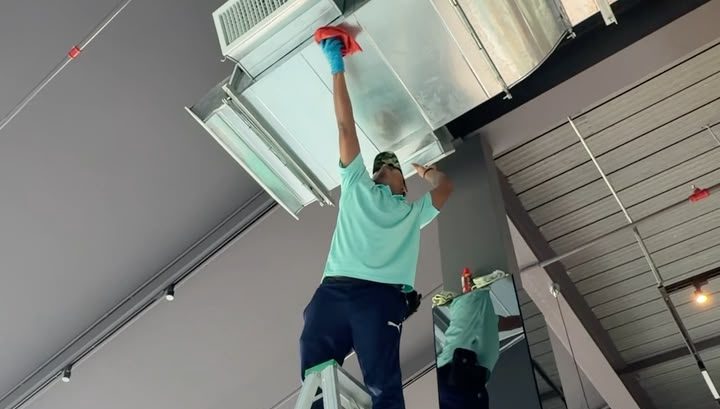 A Kean Can Cleaning Service cleaner on a step ladder wiping the outside of ceiling ductwork and a grille in a commercial interior