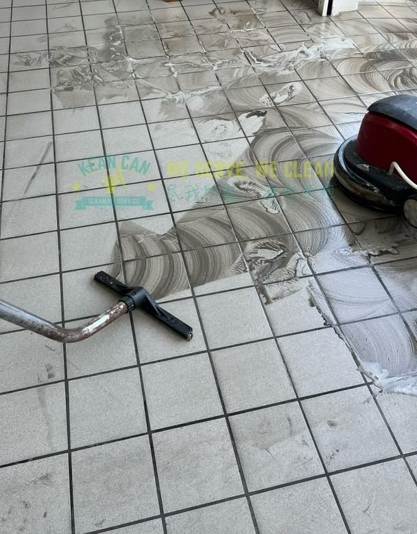 A red rotary floor machine part-way through scrubbing a grey tiled floor, the wet cleaned band curving away from the pad with the untouched tiles beside it